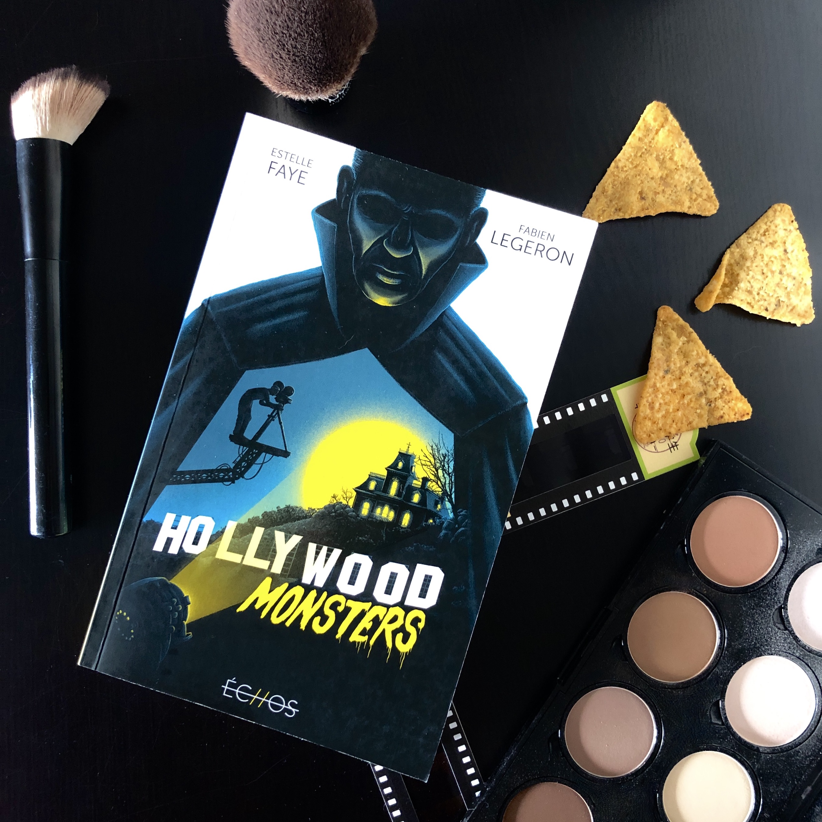 Livre Hollywood monsters posé sur une surface sombre avec des morceaux de pellicules de films, des nachos, une palette de maquillage et des pinceaux. La couverture montre une manoir victorien illuminé par un projecteur qui apparaît dans le creux d'une silhouette inquiétante avec une cape et un col haut.