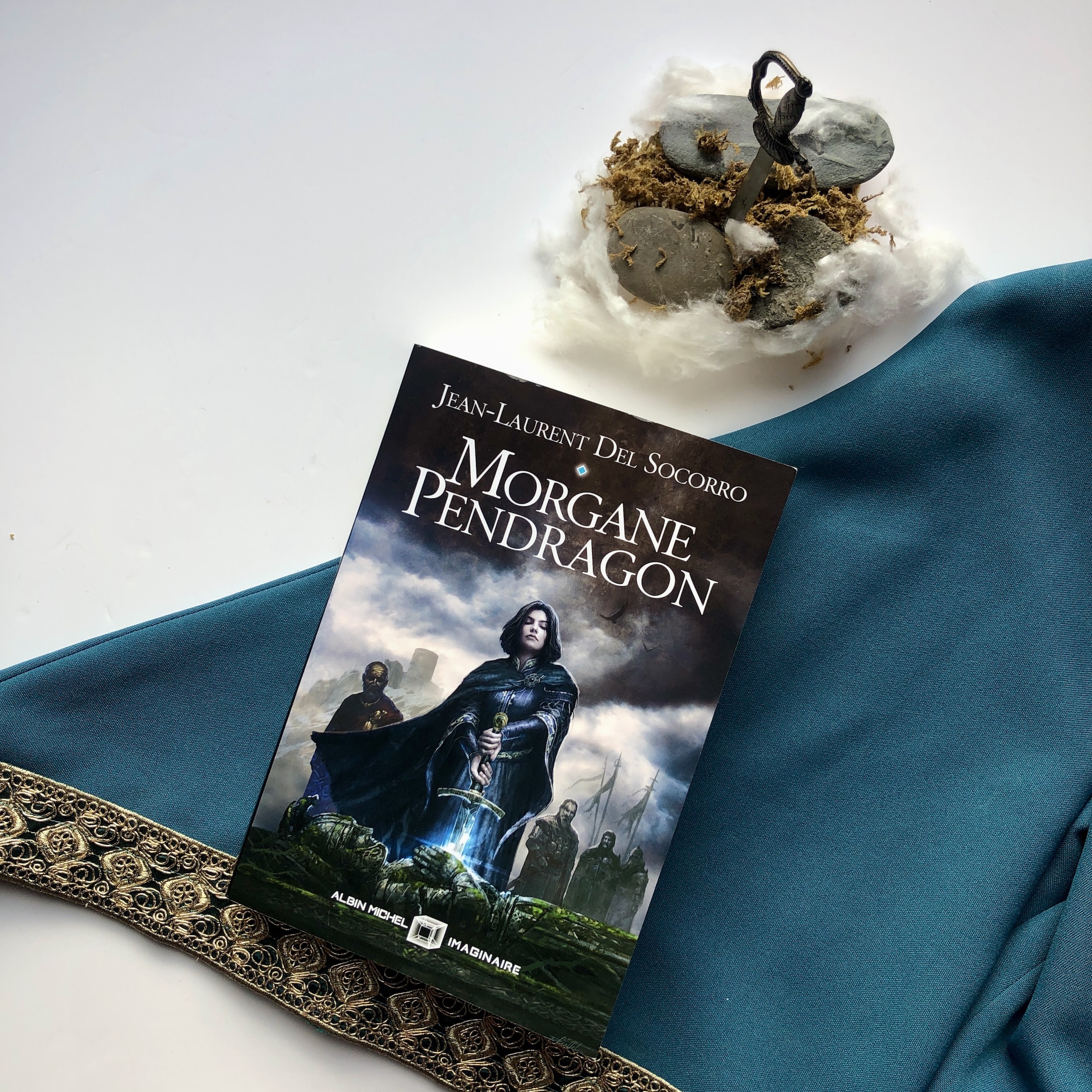 Livre Morgane Pendragon chez Albin Michel Imaginaire posé sur une manche de robe médiévale bleue à bordure dorée. Au dessus du livre, une miniature d'une îlot de mousse et de pierres dans la brume où est planté une épée. La couverture du livre montre un personnage féminin retirant une épée d'un tombeau de pierre. Derrière elle, différentes silhouettes et étendards et en fond un château dans un ciel noir et nuageux.