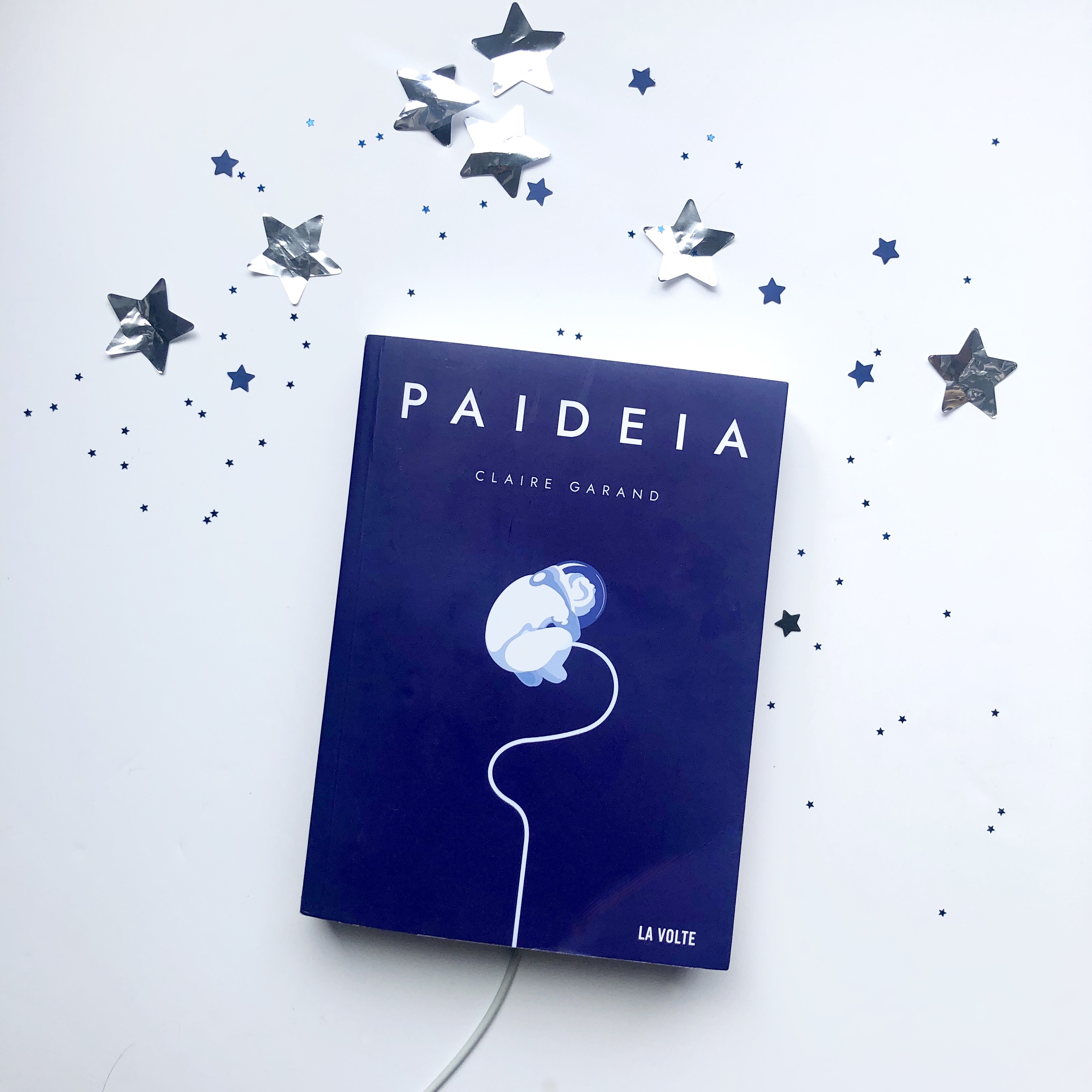 Livre Paideia de Claire Garand sur fond blanc avec des étoiles autour et un câble blanc prolongeant celui de la couverture. La couverture montre une silhouette en tenue d’astronaute en position foetale dans un fond bleu nuit. La silhouette est raccroché par un câble blanc qui sort de la couverture et qui évoque tant le câble des astronautes en EVA que le cordon ombilical.  