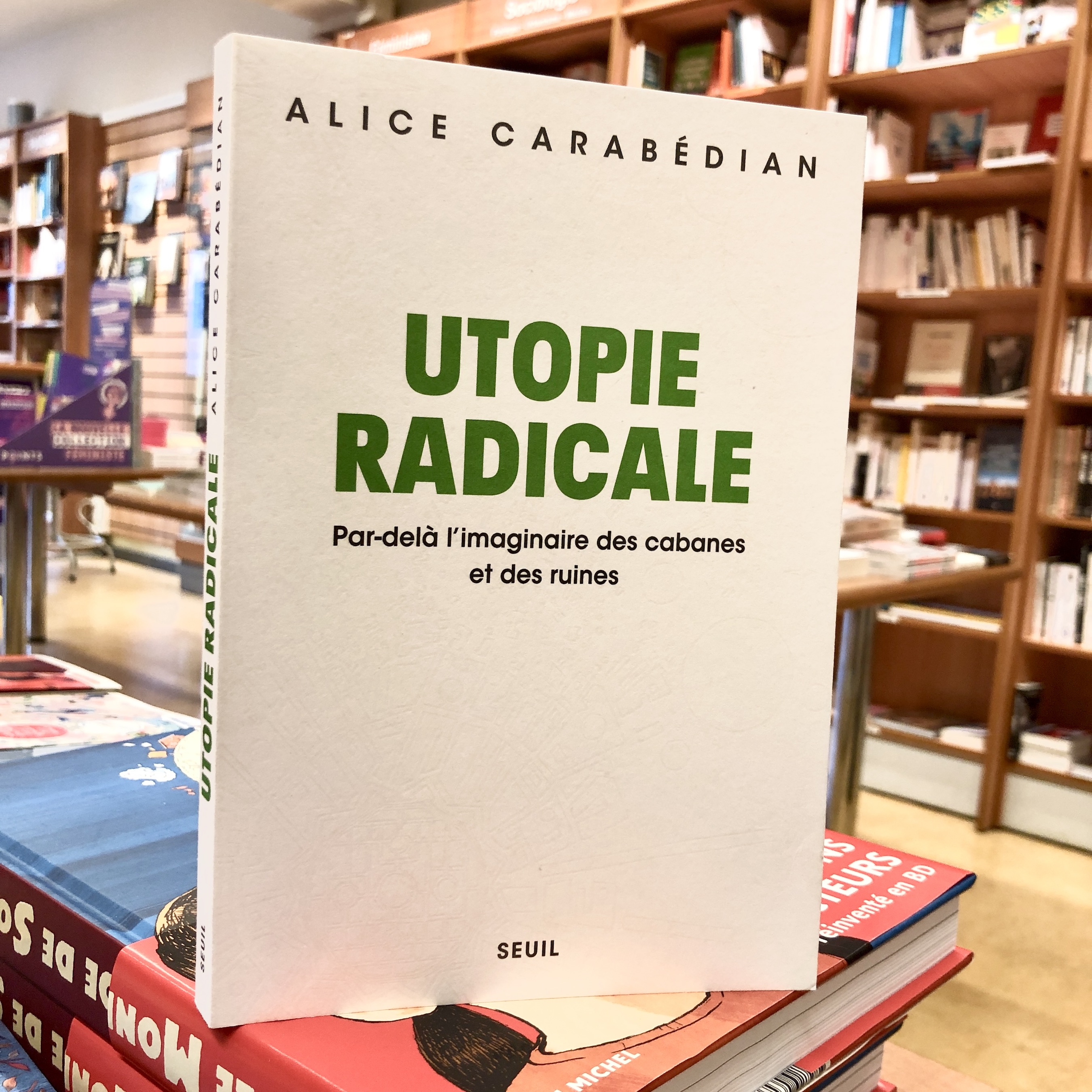 Couverture d’Utopie radicale où le titre en vers est écrit sur une couverture au fond blanc sur lequel apparaît en légère teinte grise fondue dans le blanc des éléments graphiques comme des planètes, étoiles, soleil et machines. Le livre est posé sur une table, en fond, des rayonnages de librairie. 